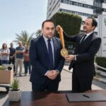 Σατιρική πολιτική εικονογράφηση με τον Νίκο Χριστοδουλίδη να απονέμει μετάλλιο στον Κώστα Κουμή ενώ συνεργάτες απομακρύνουν την υπουργική καρέκλα του