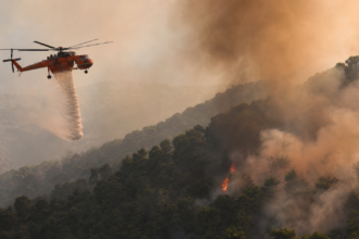 Πυροσβεστικό ελικόπτερο επιχειρεί πάνω από δασική πυρκαγιά σε συμβολική εικονογράφηση για τη σύγκρουση στην Ψάθα